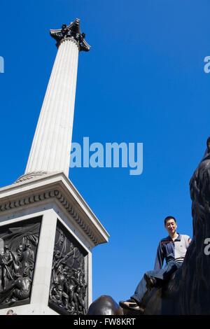 Ein Tourist aus dem Fernen Osten thront auf einer bronzenen Löwen Abgüsse in Trafalguar Square London. in den Schatten gestellt durch die Statue von Nelson auf seiner Kolumne der Tourist scheint auf Urlaub oder Ferien und schaut, um eine Fotografie genommen in einer der Hauptstädte Attraktionen oder Sehenswürdigkeiten haben. Stockfoto