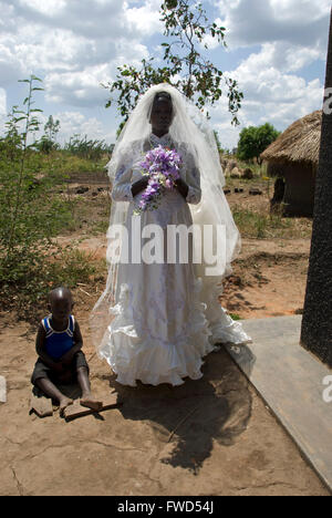 Lacekocot, Pader, Uganda. Freunde und Familie zu sammeln für die Gruppe Hochzeit von sechs Acholi Paare auf der Lacekocot-Flüchtlingslager. Eine Braut wartet geduldig vor der Kirche, ein kleiner Junge sitzt neben ihr, es gibt Lehmhütten im Hintergrund Stockfoto