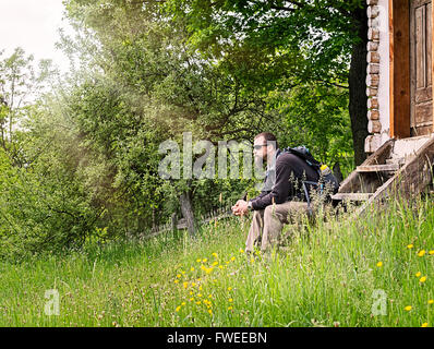 Bärtiger junger Mann mit Sonnenbrille und Rucksack ruht auf den Stufen eines alten Hauses. Stockfoto