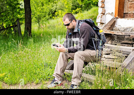 Bärtiger junger Mann mit Sonnenbrille und Rucksack ruht auf den Stufen eines alten Hauses, das Abrufen von e-Mails auf seinem Smartphone. Stockfoto