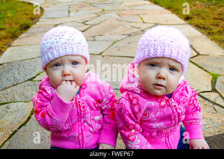 Zwei Twin Baby Mädchen sitzen auf einem steinernen Steg tragen rosa Sweatshirts und Mützen. Stockfoto