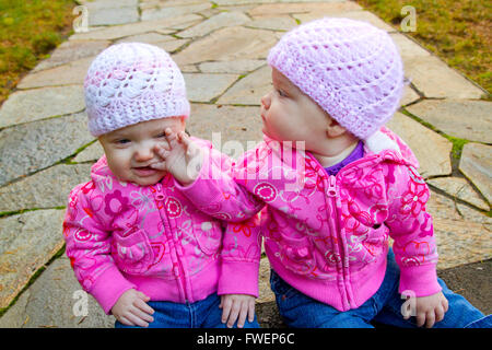 Zwei Twin Baby Mädchen sitzen auf einem steinernen Steg tragen rosa Sweatshirts und Mützen. Stockfoto