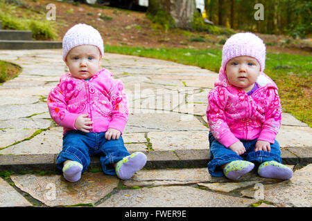 Zwei Twin Baby Mädchen sitzen auf einem steinernen Steg tragen rosa Sweatshirts und Mützen. Stockfoto