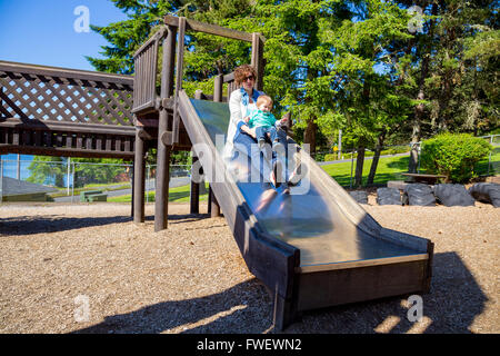 Eine Mutter und ihr Sohn rutschen ein Metall schieben an einen Spielplatz im Freien in einem Park. Stockfoto