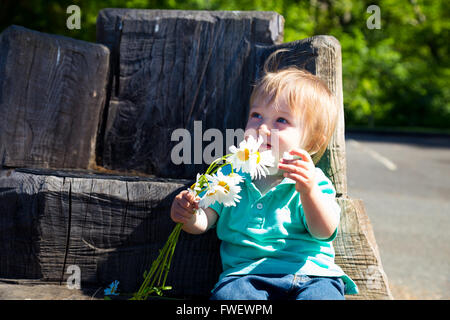 Ein kleiner Junge spielt mit einigen Blumen sitzend auf einer Hand geschnitzt Sitz aus einem alten stumpf gemacht. Stockfoto