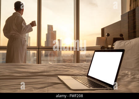 Konzentrieren Sie sich auf Laptop-Computer mit Kopie Speicherplatz leeren Bildschirm auf dem Bett. Jungunternehmer im weißen Bademantel am Fenster stehen Stockfoto