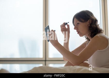 Lifestyle Portrait junge Frau sitzt auf dem Bett und setzen auf Mascara nach dem Aufstehen. Kaukasische weibliches model Stockfoto