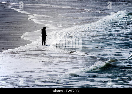 Eine Frau in der Silhouette und in einem Abstand allein stehend auf den Fistral Beach in Newquay, Cornwall gesehen. Stockfoto