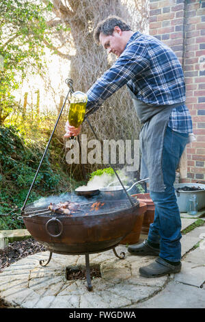 Ein Mann Kochen Braten Wildgeflügel über dem offenen Feuer gießen Öl aus einer Flasche auf das Fleisch. Stockfoto