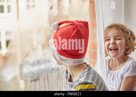 Weihnachtsmorgen in einem Einfamilienhaus. Ein Junge in eine Weihnachtsmütze aus einem Schlafzimmerfenster und seine Schwester neben ihm lachend. Stockfoto