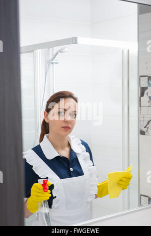 Einzelne niedliche junge Magd mit ernsten Ausdruck Blick in Spiegel halten Sie klare Sprühflasche und Tuch im gelben behandschuhten Händen. Stockfoto