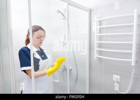 Magd, Hotelangestellter oder Haushälterin Reinigung ein Bad, das Glas der Duschkabine abwischen, da sie im Inneren befindet sich in einer weißen Schürze. Stockfoto