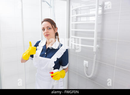 Überzeugt junge weibliche Hausangestellte im blauen Kleid und weiße Schürze mit Daumen hoch Geste gedrückter Sprühflasche neben Duschkabine im Badezimmer stehen. Stockfoto
