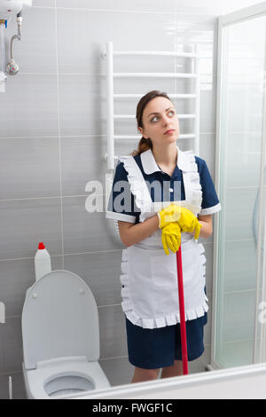 Nachdenkliche junge Magd oder Haushälterin eine Pause von ihrer Arbeit stehen, stützte sich auf ihre Mop in die Luft starren Stockfoto