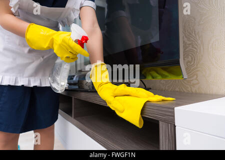 Magd Reinigung einen TV-Schrank Besprühen mit Reinigungsmittel vor dem Abwischen mit einem Tuch hautnah auf ihre behandschuhten Hände Stockfoto