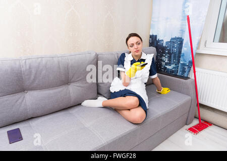 Faul, Haushälterin oder Dienstmädchen in einheitlichen eine Pause aufstellen ihre Füße auf dem Sofa wie sie eine Fernsehsendung wählt ansehen Stockfoto