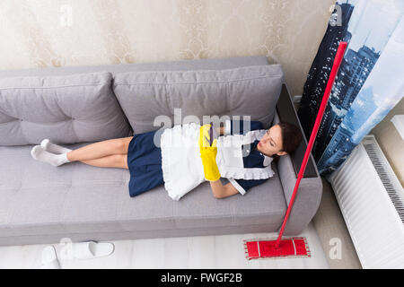 Faule Arbeit schüchtern Haushälterin in uniform eine Pause mit ihren Mopp neben ihrer Überprüfung ihres Mobiltelefons, Blick von oben auf dem Rücken auf einem Sofa liegend. Stockfoto