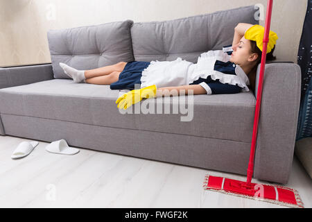 Entspannen Sie sich auf den Job auf einer Couch in ihrem einheitlichen ein Nickerchen mit ihren Mopp neben erschöpft Haushälterin Stockfoto