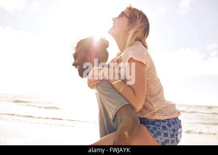 Seite Ansicht Porträt junger Mann mit seiner Freundin auf dem Rücken am Strand. Mann Huckepack Freundin am Meeresufer auf Sonne Stockfoto