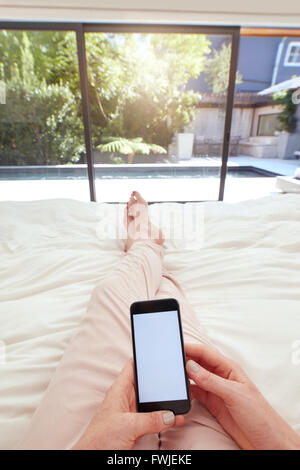 POV Aufnahme einer Frau mit Handy auf dem Bett liegend. Menschliche Hand halten Smartphone mit leeren Bildschirm im Schlafzimmer. Stockfoto