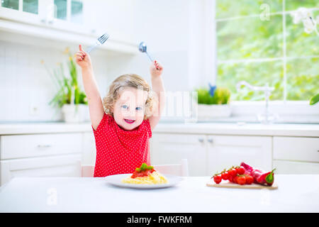 Niedliche lockige lachende Kleinkind Mädchen in ein rotes Hemd mit Gabel und Löffel essen Spaghetti mit Tomatensauce und Gemüse spielen Stockfoto
