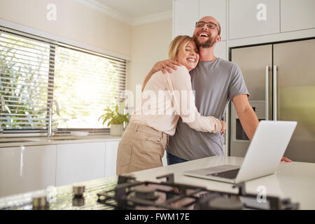 Porträt der glückliche Frau umarmte ihren Mann in der Küche. Liebespaar mit Laptop am Küchentisch zu Hause. Stockfoto