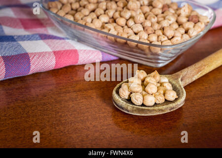 Ungekochte Kichererbsen in alten Holzlöffel mit Glasschüssel mit Kichererbsen und Küche Rag im Hintergrund. Textfreiraum Stockfoto