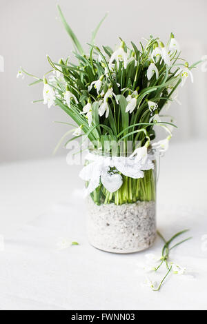 Schönen Schneeglöckchen Blüten in eine transparente Vase mit Spitzenband und ein kleines Herz auf einem weißen Tisch Stockfoto