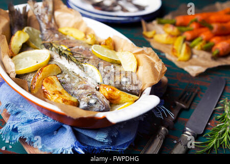 Ein Ofen gebratene Forellen in einem hohen Topf auf einem rustikalen Tisch. Mit Karotten und Kartoffeln auf der Seite. Stockfoto