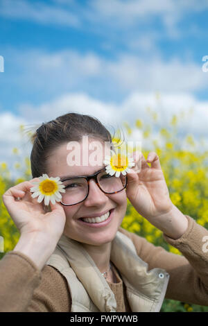 Lächelnde junge Frau hält ein paar Gänseblümchen Blumen auf beiden Seiten des Gesichts unter blauem Himmel mit gelben Wildblumen in der Stockfoto