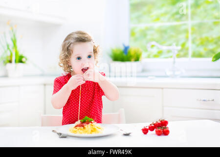 Niedliche lockige lachende Kleinkind Mädchen in ein rotes Hemd mit Gabel und Löffel essen Spaghetti mit Tomatensauce und Gemüse spielen Stockfoto