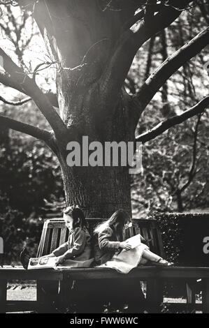 zwei kleine Mädchen Schwestern sitzen auf Bank unter Baum und lesen Sie Bücher Stockfoto