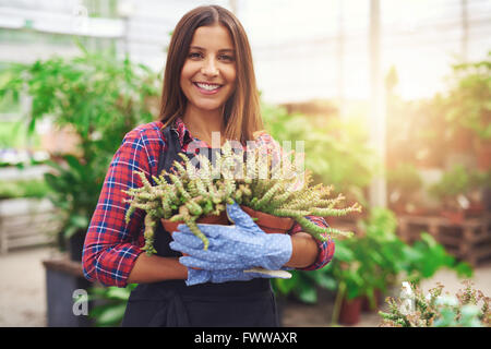 Attraktive erfolgreiche Baumschule Inhaber in Ihrem Gewächshaus Holding eine Topfpflanze in ihre Hände, als sie verleiht der Kamera eine freundliche smi Stockfoto