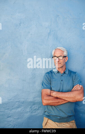 Porträt von einem Reifen kaukasischen Mann mit Brille stehend mit seine Arme verschränkt vor blauem Hintergrund. Mitte erwachsenen Mannes auf der Suche nach aw Stockfoto