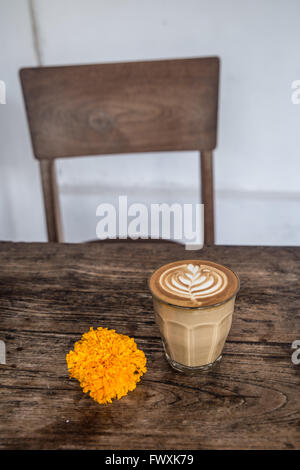 Cafe Latte Glas mit Latte Art und Orange Blume auf hölzernen Tisch und Stuhl im Hintergrund Stockfoto