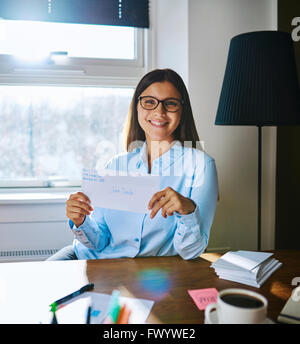 Glückliche single-Frau in Gläsern und leichte blaue Bluse Holding Umschlag mit handschriftlichen Adresse hinter Schreibtisch sitzend Stockfoto