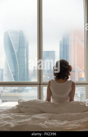 Rückansicht des junge Frau sitzt auf ungemachten Bett nach dem Aufwachen und mit Blick auf Stadt-Landschaft im Fenster. Weibliches Model entspannend Stockfoto