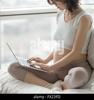 Schwangere Frau sitzt mit gekreuzten Beinen in bequemen Haltung auf Bett mit Laptop. Zukünftige Mama werdende Kind Schlafzimmer zu Hause entspannen Stockfoto