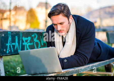 Schöne elegante Geschäftsmann sitzt auf einer Holzbank, arbeiten im Freien in einem städtischen Park, die Eingabe von Informationen auf seinem Laptop comp Stockfoto