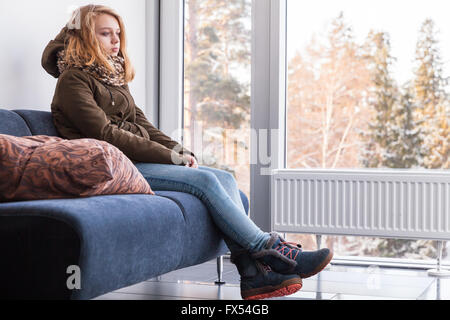 Schöne blonde kaukasische Teenager-Mädchen in warme Kleidung, die sitzt auf dem Sofa neben dem Fenster im Flur Stockfoto