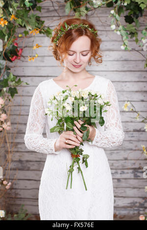 Junge rothaarige Braut in einem grünen Kranz steht unter dem Bogen der Herbstpflanzen und halten einen weißen Blumenstrauß Stockfoto