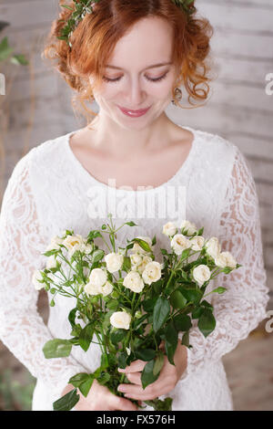 Porträt von eine junge rothaarige Braut in einem einfachen Spitzenkleid mit einem Bouquet aus weißen Rosen. Stockfoto