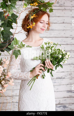 Junge rothaarige Braut in einem einfachen weißen Spitzenkleid hält ein Bouquet von kleinen weißen Rosen. Stockfoto