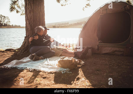 Porträt von senior Liebespaar camping am Ufer eines Sees an einem schönen Sommertag. Sie sitzen unter einem Baum vor dem Zelt, Stockfoto
