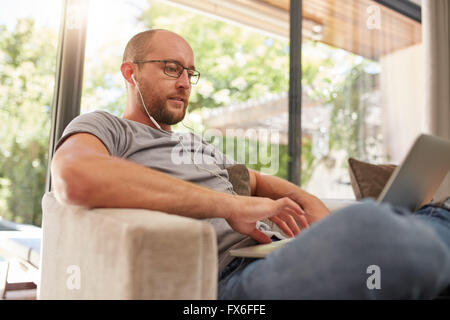 Schöner reifer Mann mit seinem Laptop beim Entspannen zu Hause, er ist mit Ohrhörer sitzen auf dem Sofa zu Hause. Stockfoto