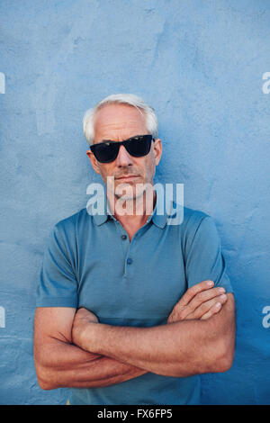 Porträt von stilvollen reifer Mann mit Sonnenbrille steht mit seinen Armen gekreuzt vor blauem Hintergrund Stockfoto