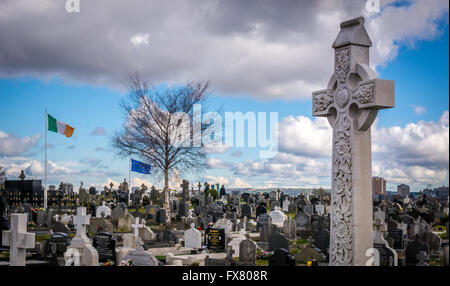 Die irische Trikolore und Starry Pflug fliegen im Hintergrund ein keltisches Kreuz und Gräber auf dem Milltown-Friedhof in Belfast. Stockfoto