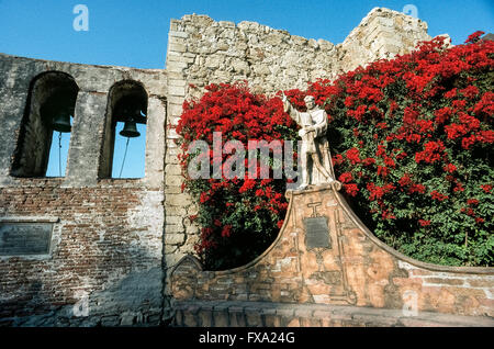 Leuchtende Bougainvillea Blumen umgeben eine Statue des spanischen Missionar Junipero Serra umarmt einen jungen, ein 1914 Denkmal des amerikanischen Künstlers Tole Van Rensaalar im Hof der Mission San Juan Capistrano in Orange County, Kalifornien, USA. Der sagenumwobene Franziskanermönch gründete die Mission im Jahr 1776, der siebte von 21 California Missionen nach oben und unten den Staat gegründet, um Spaniens Gebiet zu erweitern und die Indianer zum Christentum zu bekehren. Pater Serra wurde heilig gesprochen, als ein katholischer Heiliger von Papst Francis in 2015. Stockfoto