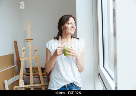 Lächelnd hübsche Frau Künstler Kaffee trinken in der Nähe des Fensters in Werkstatt Stockfoto