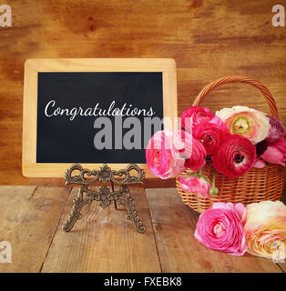 Rosa Blumen neben Tafel mit Satz: Wir gratulieren, Holztisch. Stockfoto